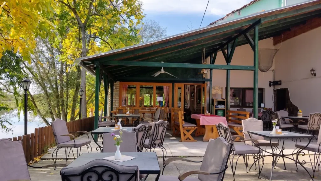 The riverside outdoor terrace of Čarda na Jami in Begečka Jama Nature Park, featuring wrought-iron chairs and a green canopy overlooking the water.